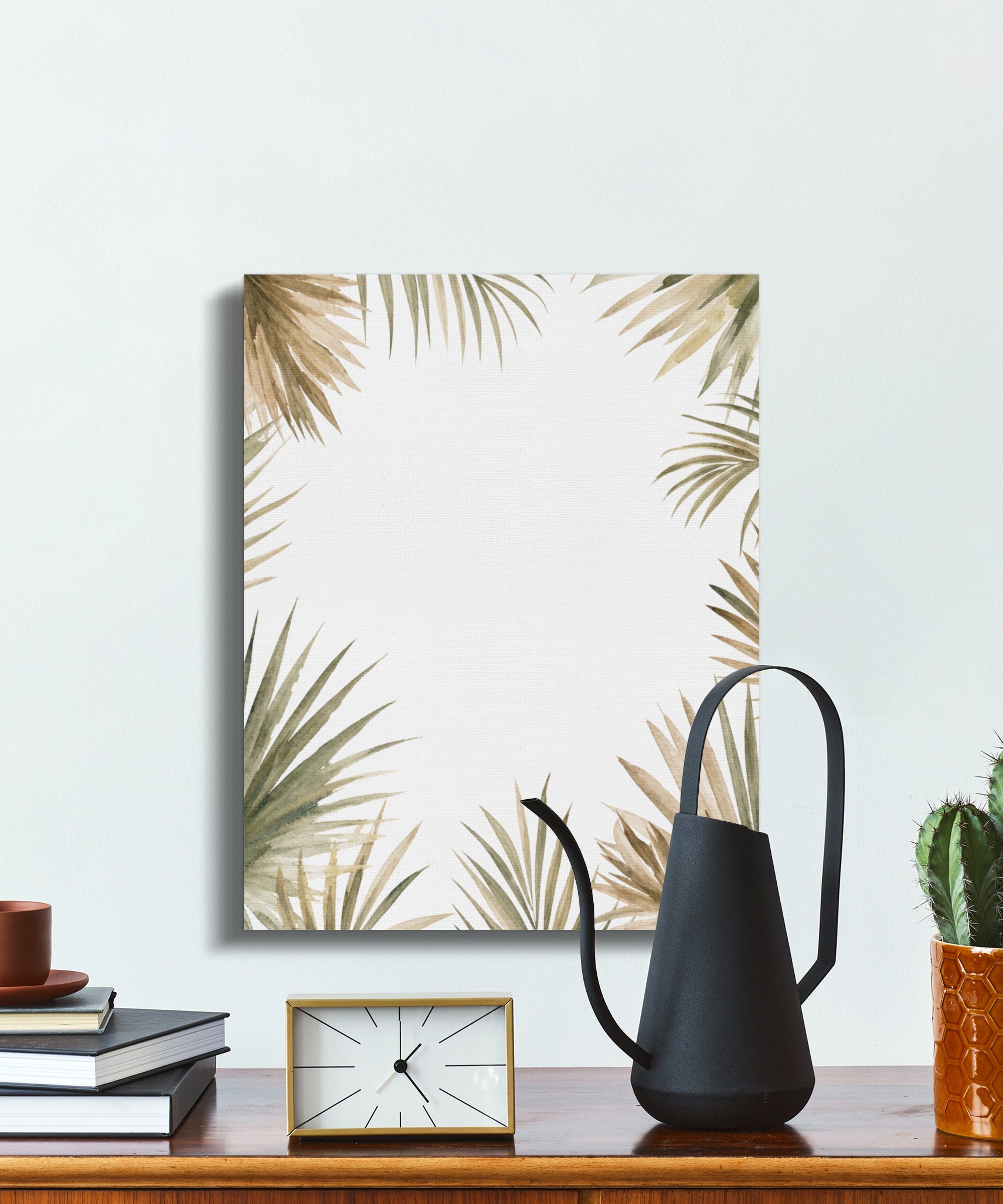 Canvas art with palm leaves and a black watering can on a wooden table.